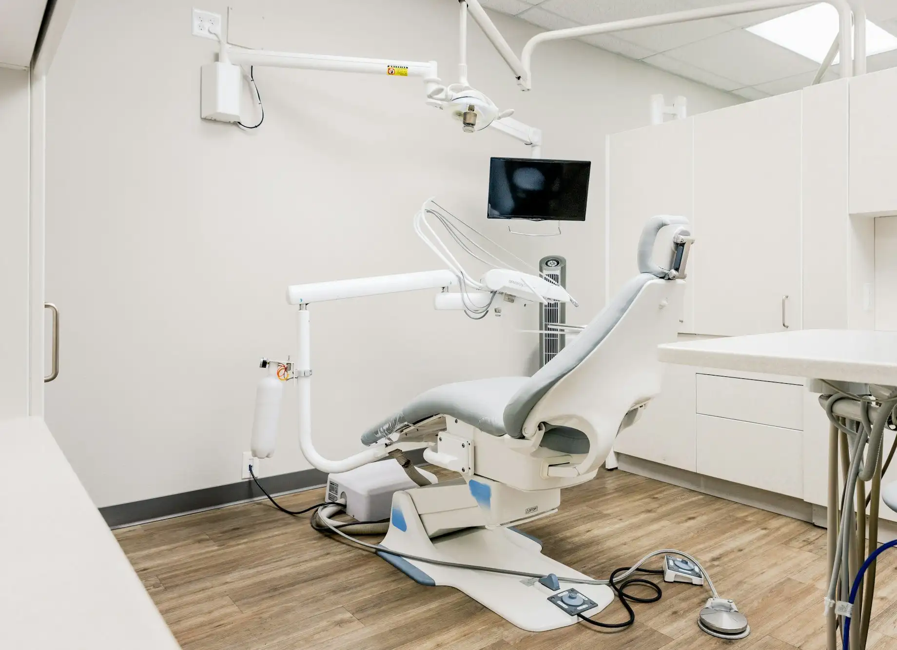 A bright dental treatment room with a pale blue chair, a monitor on an arm, white cabinets and a wood-look floor