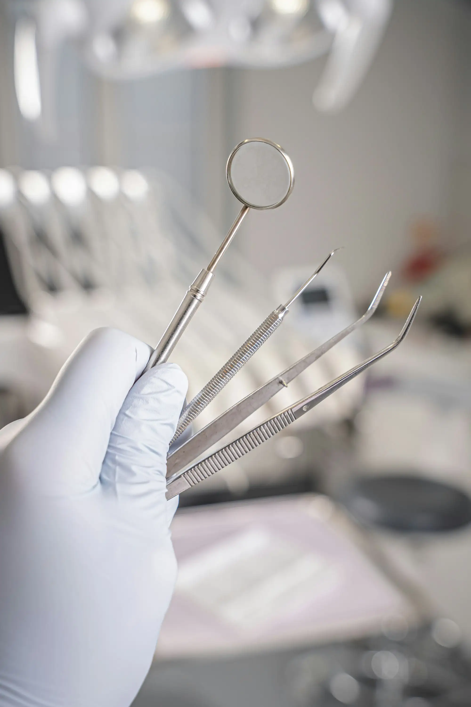A gloved hand holding a dental mirror, a probe and two pairs of tweezers in a bright treatment room