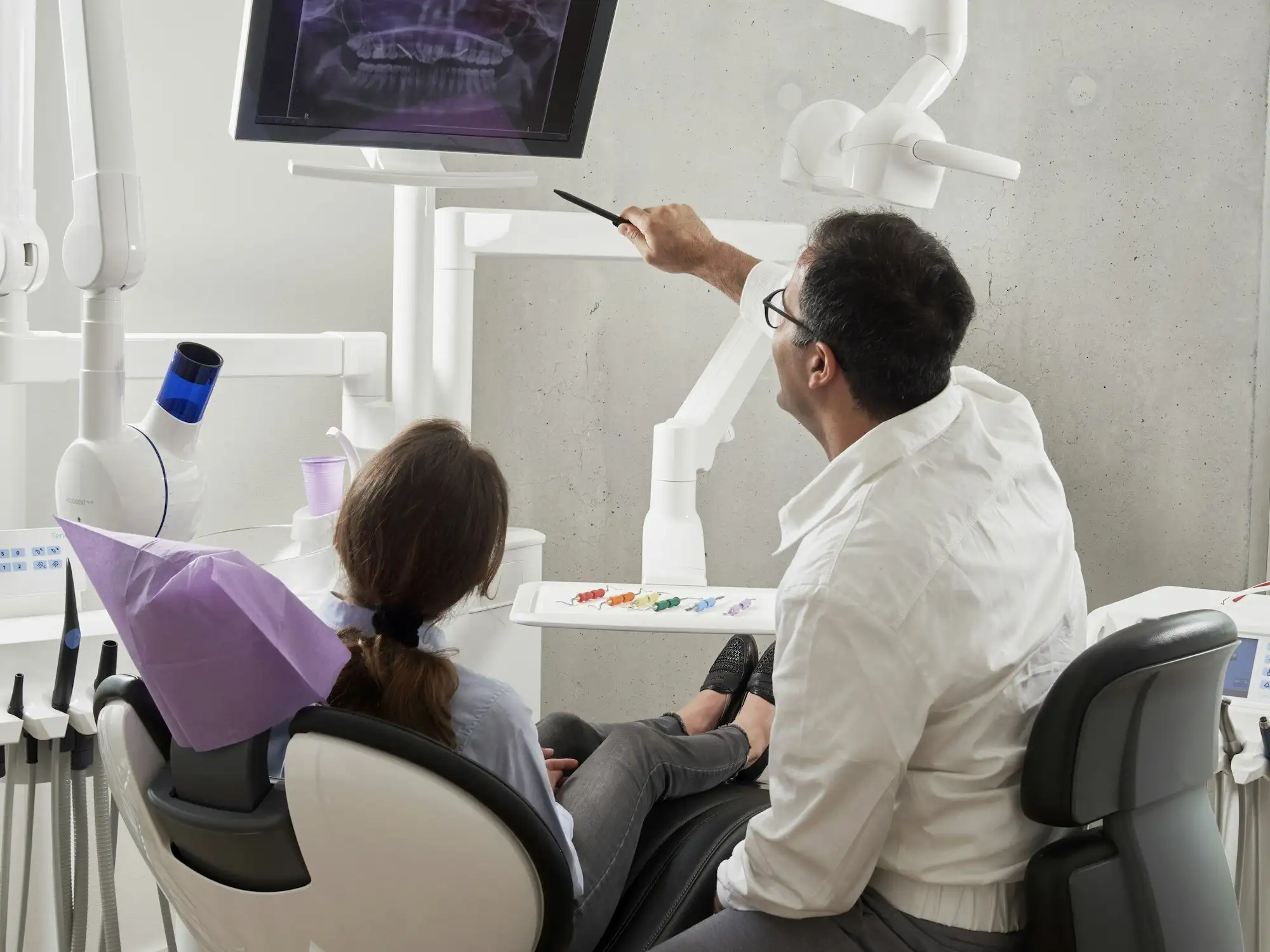 A dentist pointing at a dental X-ray on a screen while a patient in the chair looks on