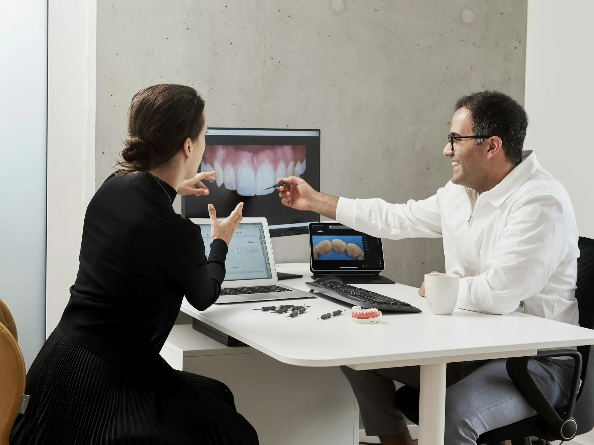 A dentist and a patient at a desk looking at a close-up of front teeth on a large screen