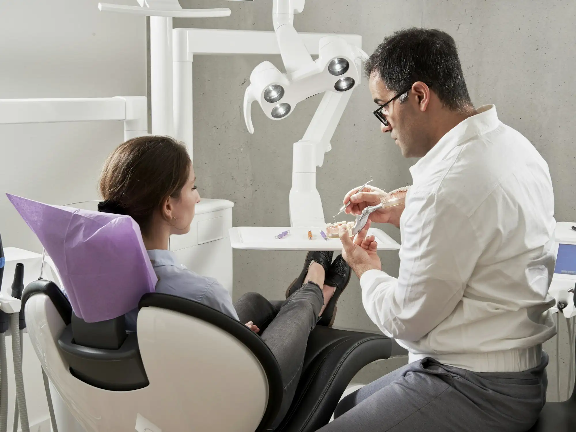 A dentist in a white shirt explaining a dental model to a patient sitting in the treatment chair