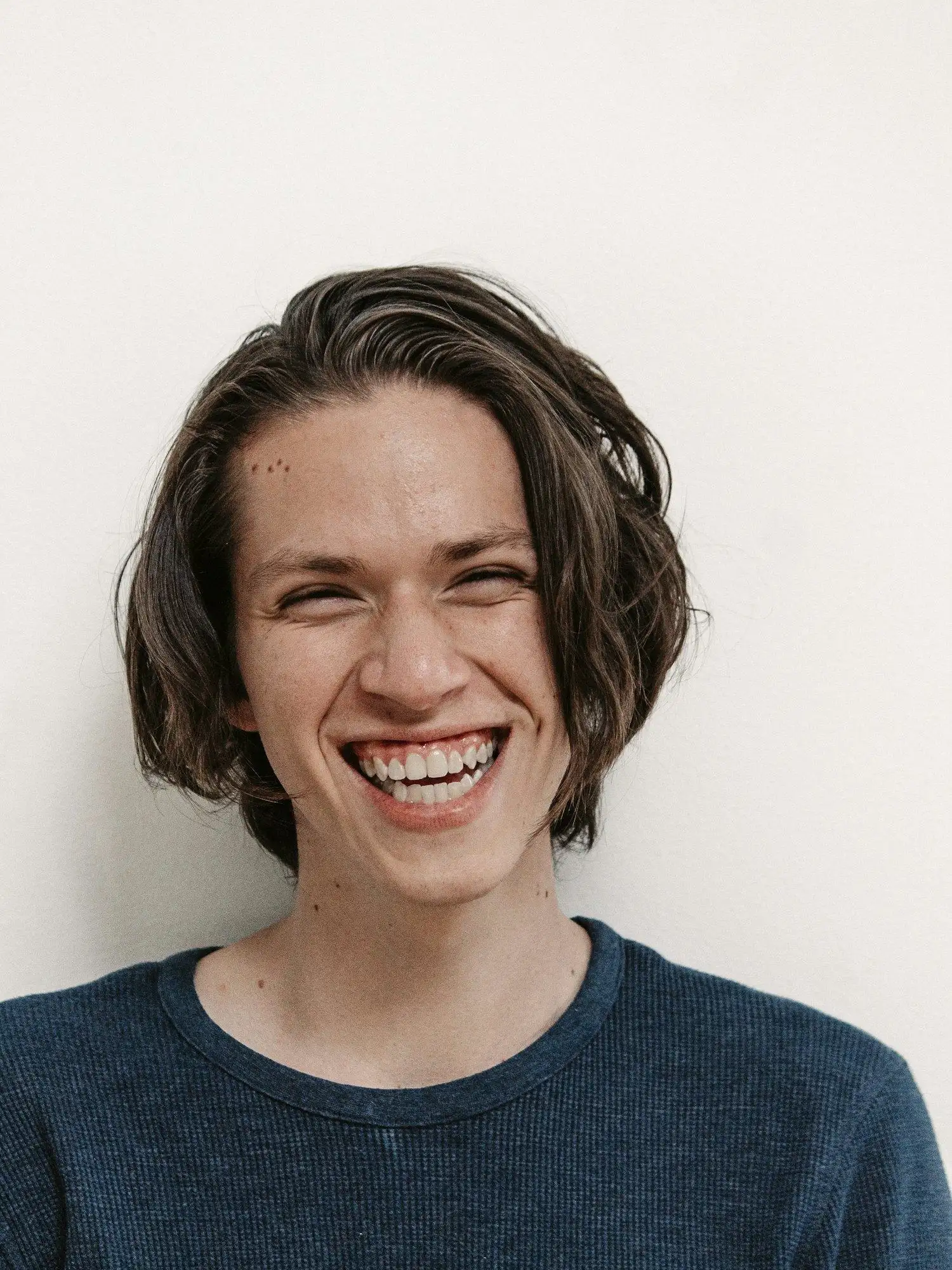A young man with shoulder-length hair laughing against a plain white wall